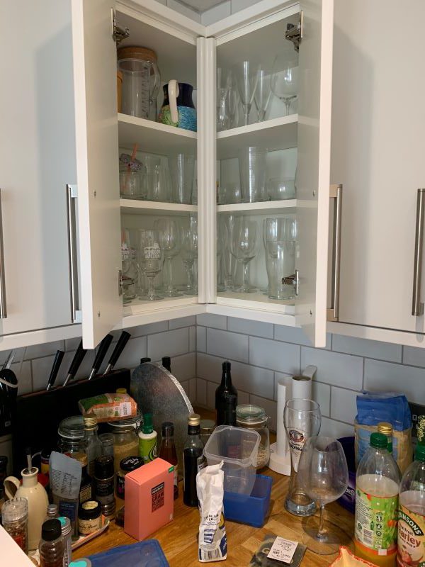 kitchen 2 Mid-organisation of a kitchen cupboard, there are plates, bowls and other items on the kitchen counter below, as well as a large quantity of bottles on a cupboard shelf, an empty cupboard shelf, and a pile of miscellaneous containers on another cupboard shelf